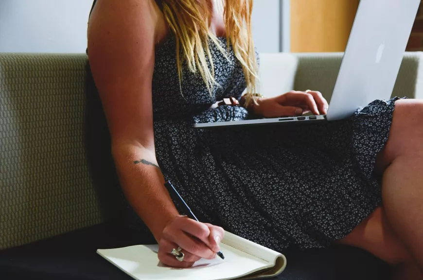 A person writing in a notebook with a laptop, sitting on a couch