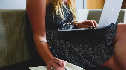 A person writing in a notebook with a laptop, sitting on a couch